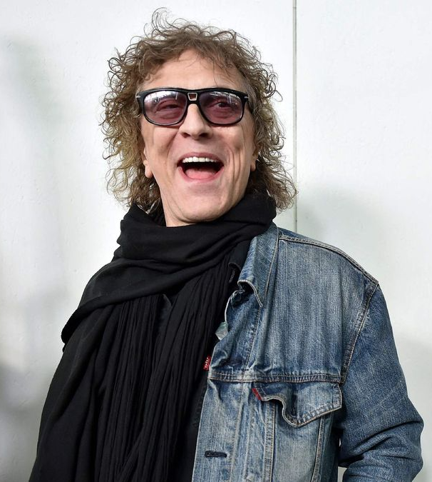 Mick Rock laughing, wearing dark glasses, a black scarf and a denim jacket.